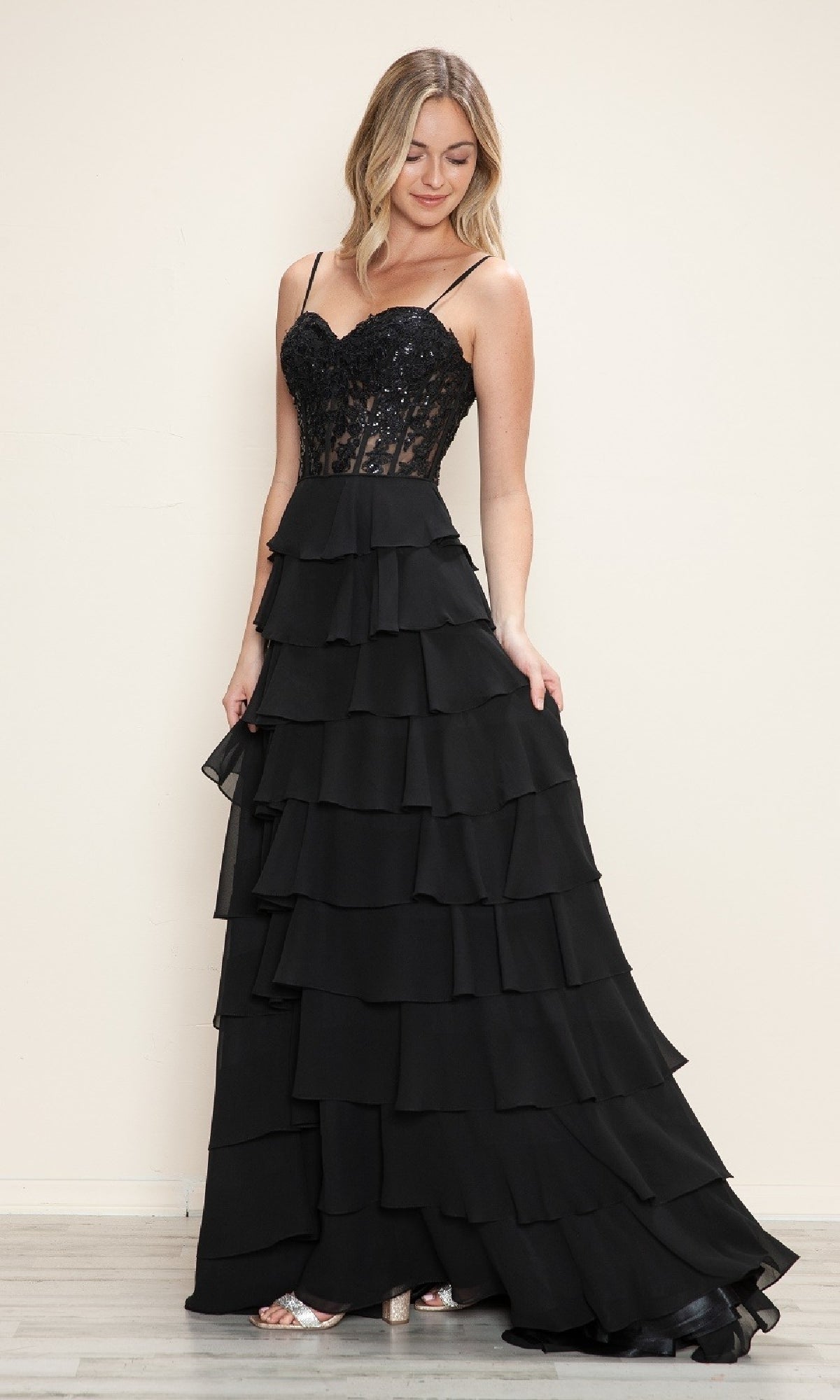 A woman wears the Poly USA 9698 Long Tiered Corset Prom Dress, a black gown with a sequined corset bodice, thin straps, and a multi-layered tiered skirt, standing before a plain light-colored background.